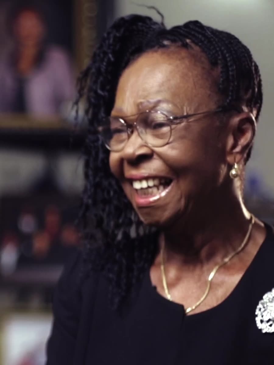 Mrs Atiomo at home, laughing, family photographs on the shelves behind her